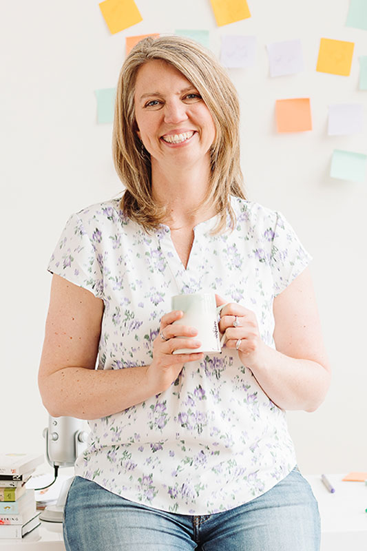 A woman, Brett Macdonald, sitting on top of her desk in front of a wall covered in sticky notes. She is holding a mug and smiling at the camera.